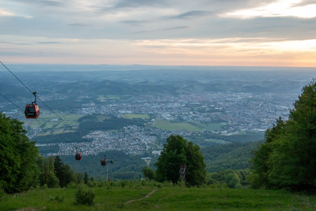 Gondolske kabine nad zelenim pobočjem s pogledom na Maribor.