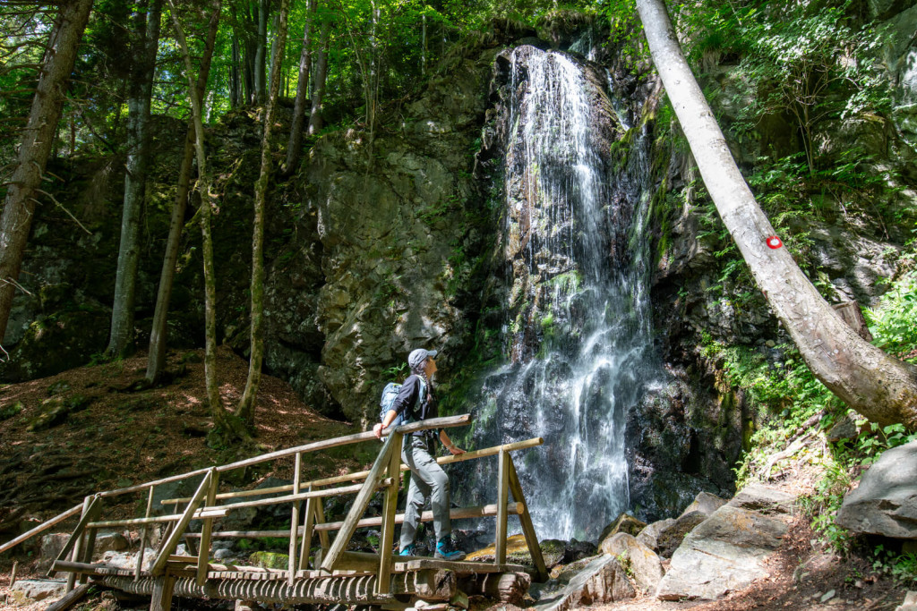 Hiker on a wooden footbridge in front of Skalca Waterfall in the green Pohorje forest.