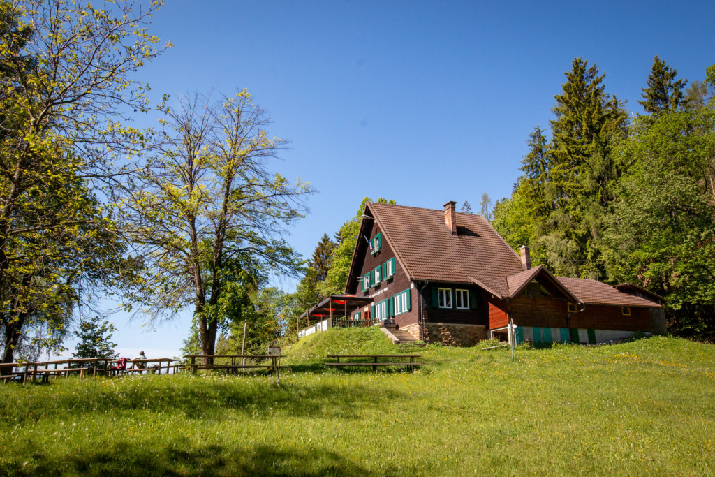 Koča na Žavcarjevem vrhu on a sunny meadow surrounded by green forest.